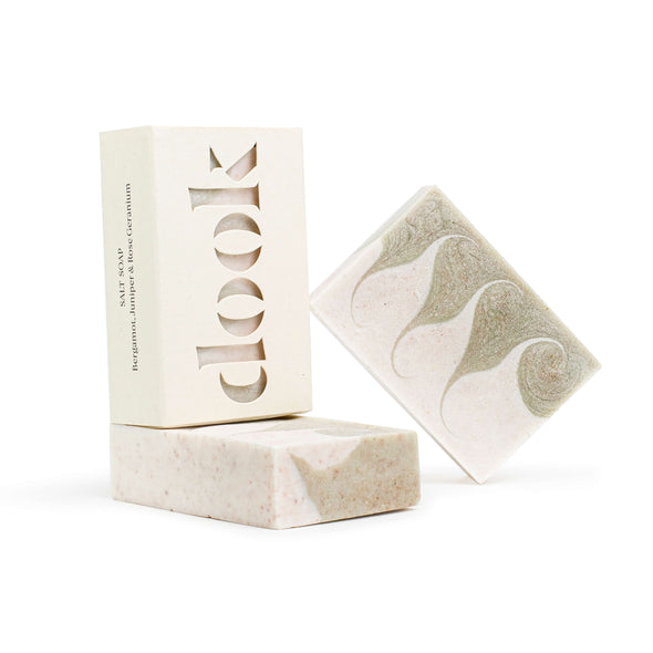 Two rectangular soaps with a white and pale green wave pattern. One lies flat and the other rests diagonally against it. A soap encased in its cardboard packaging sits atop the soap lying flat. The brand name 'dook' is cut out on the front of the packaging box.