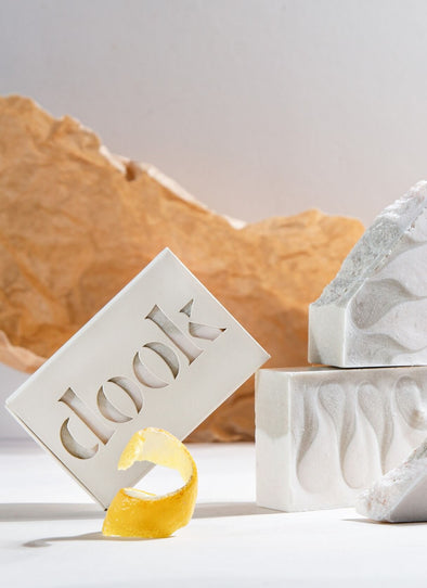 Three rectangular soap bars with a white and pale green wave pattern. One is encased in cardboard packaging with the brand name 'dook' cut out on the front. A curled lemon rind is in front and a styled piece of crumpled brown paper provides a backdrop.