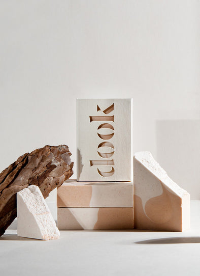 Four rectangular soap bars with a white and pinky orange wave pattern. One is encased in cardboard packaging with the brand name 'dook' cut out on the front. The soap bars are styled on top of and next to each other, alongside a piece of dried wood.