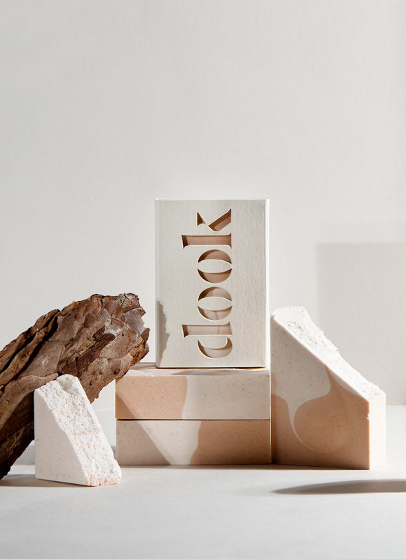 Four rectangular soap bars with a white and pinky orange wave pattern. One is encased in cardboard packaging with the brand name 'dook' cut out on the front. The soap bars are styled on top of and next to each other, alongside a piece of dried wood.
