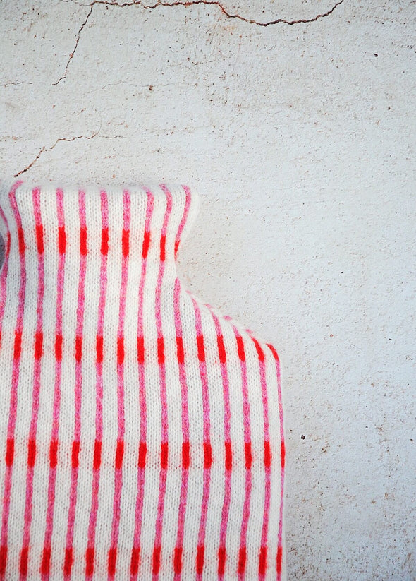 Close up corner of a white knitted lambswool hot water bottle with thin vertical stripes in red and pink.