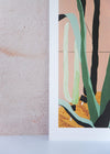 Close up of a corner of an art print by Rachel Victoria Hillis. Depicting various green cacti on a pale pink background and sandy floor.