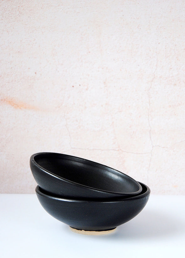 Two footed bowls stacked on top of each other. Handmade by Dor and Tan using local Cornish clay, and finished in a soft matte black glaze. Each bowl is five centimetres high and sixteen centimetres wide.