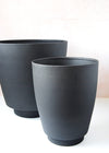 Two black metal planters, the smaller one is in front, to the side, of the larger one. The small one is 25cm tall and 21cm wide. The large is 31cm tall and 30.5cm wide.