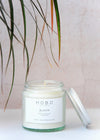 Aromatherapy travel candle, handmade by HoBo Soy Candles using one hundred percent essential oils. The label shows the scent as being rose geranium and lavender. The candle is presented in a one hundred and twenty millilitre recycled clear glass jar with a metal lid leaning against the side. The jar is six centimetres high and five point five centimetres wide.