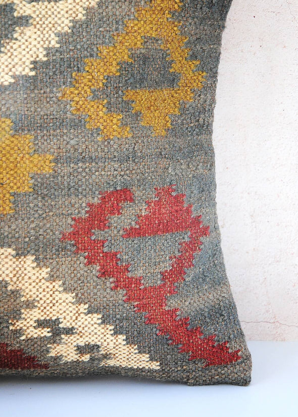 Close up corner of a blue cushion with a yellow, red and cream pattern. Made from wool and jute with a one hundred percent cotton back with zip closure. Measures forty five centimetres by forty five centimetres.