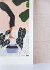 Close up corner of an art print by Rachel Victoria Hillis. Depicting a woman doing a yoga pose surrounded by green leaves and plants, on a pale pink background and white floor with black polka dots.