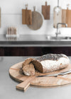 Round wooden bread board resting on a kitchen work surface. A partially sliced loaf of bread and a silver metal knife rest on top.