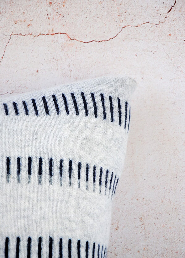 Close up corner of a grey knitted lambswool cushion with a navy blue dash pattern. Height forty five centimetres, width forty five centimetres.