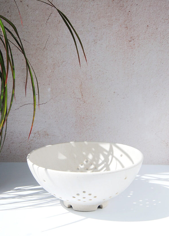Ceramic berry bowl in a white glaze. A few plant fronds can be seen in the top left corner casting shadows over the bowl. The bowl is 9.5cm high and 21.3cm wide at the top.