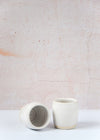 Pair of espresso cups handmade by Dor and Tan using local Cornish clay, one standing upright and one lying on its side. Each cup has a white glaze, no handle and is approximately six centimetres tall and 6 centimetres wide.