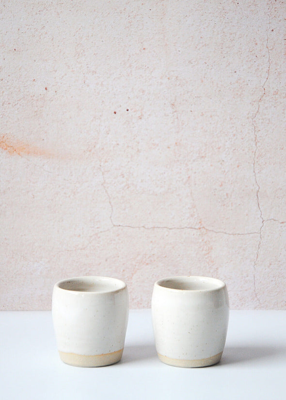 Pair of espresso cups handmade by Dor and Tan using local Cornish clay. Each cup has a white glaze, no handle and is approximately six centimetres tall and six centimetres wide.