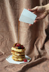 Hand pouring sauce from a geometric milk jug over a stacked pile of pancakes with fruit topping.