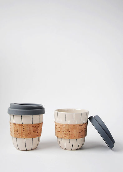 Two reusable ceramic coffee cups sitting side by side. They both have a cream base and are hand decorated - one with dark grey vertical stripes and the other with a dark grey dash pattern. They both have cork sleeves and grey blue silicone lids. The cup on the left is open with its lid resting against the side of it. Each cup is 10cm high (without lid), and 8.5cm wide (at top)