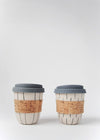 Two reusable ceramic coffee cups sitting side by side. They both have a cream base and are hand decorated - one with dark grey vertical stripes and the other with a dark grey dash pattern. They both have cork sleeves and grey blue silicone lids. Each cup is 10cm high (without lid), and 8.5cm wide (at top)