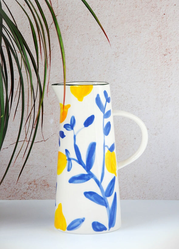 Ceramic jug with a cream base featuring a painted pattern of yellow lemons and blue twigs with a green rim at the top and a handle on the side. Height twenty centimetres, width nine centimetres at bottom.