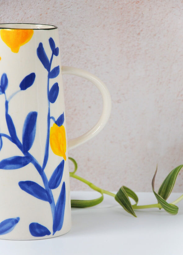 Close up section of a ceramic jug with a cream base featuring a painted pattern of yellow lemons and blue twigs with a green rim at the top and a handle on the side. Height twenty centimetres, width nine centimetres at bottom.