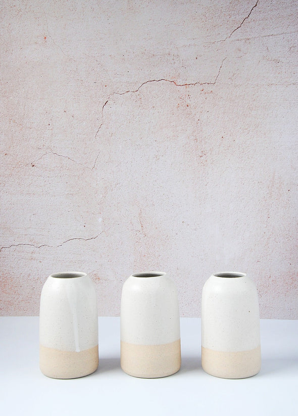 Three freckled ceramic bottle vases lined side by side in a row. The top two thirds of each vase are covered in a white glaze while the bottom is left raw. Each vase is 13cm high and 8cm wide (at base).