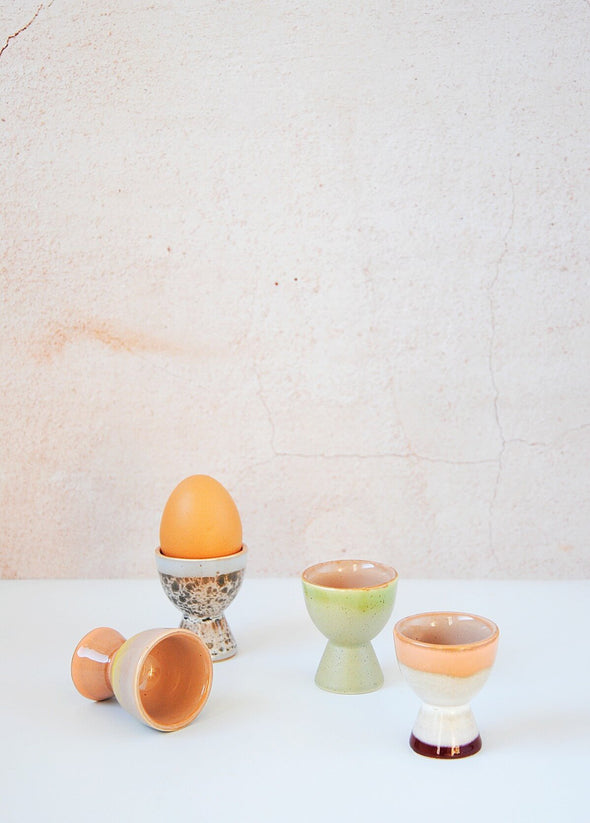 Four ceramic egg cups in various glazes of greens, blues and pinks. One egg cup contains an egg and one lies on its side. The other two are upright.