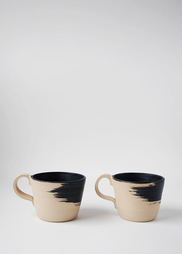 Two stoneware mugs side by side.They have a raw finish and a matt black brush stroke design on the exterior. The interior of the mug is painted in a matt black glaze.