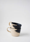 Two stoneware mugs in a stack. They have a raw finish and a matt black brush stroke design on the exterior. The interior of the mug is painted in a matt black glaze.