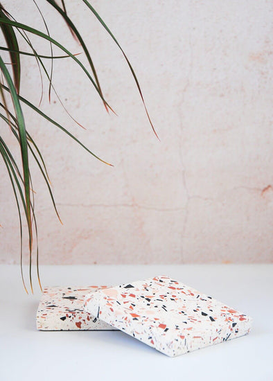 Two terrazzo square coasters handmade by Katie Gillies using jesmonite. They have a white base and brown, black and pink chips. They are each ten centimetres by ten centimetres.