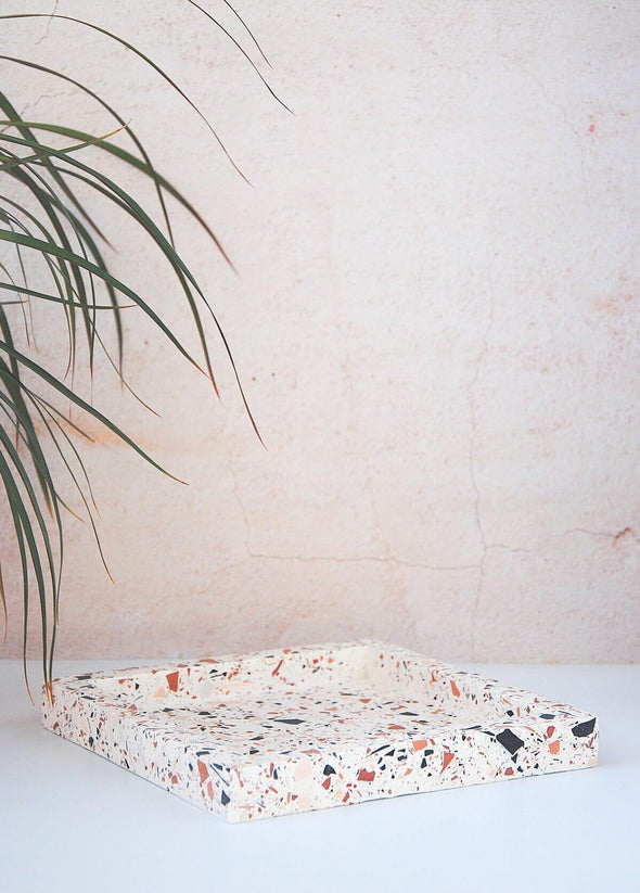 Terrazzo square tray handmade by Katie Gillies using jesmonite. It has a white base and brown, pink and black chips. It is twenty centimetres by twenty centimetres and is two centimetres thick.