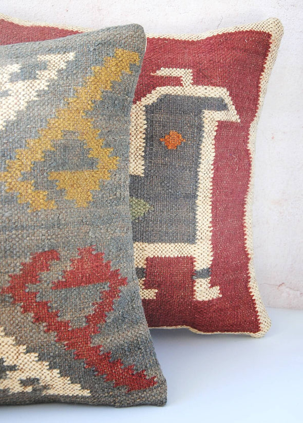 Close up of a pair of cushions made from wool and jute. The cushion behind is red with a blue and cream pattern, the cushion in front is blue with a yellow, red and cream pattern. Each cushion measures forty five centimetres by forty five centimetres.