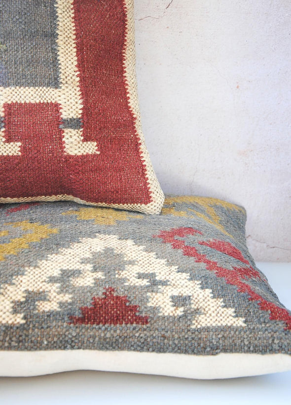 Close up of a pair of cushions made from wool and jute with a one hundred percent cotton back featuring a zip fastening. The cushion on top is red with a blue and cream pattern, the cushion underneath is blue with a yellow, red and cream pattern. Each cushion measures forty five centimetres by forty five centimetres.