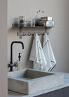 A concrete square sink and black tap. On the wall behind is a metal shelf with hooks below. On the shelf are assorted drinking glasses and two metal storage tins. Hanging from the hooks are two kitchen towels. They are white with a dark green stripe and check design.