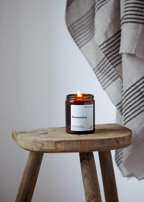Scented candle in a brown apothecary inspired jar, made by Earl of East London using only natural soy wax. The candle is placed on a wooden stool with fabric hanging in the background. The label of the candle shows the name as being Elementary. The jar has a capacity of one hundred and seventy millilitres and is eight centimetres tall and six point five centimetres wide