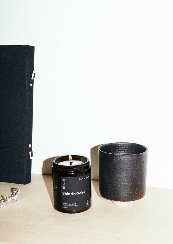 Scented candle in a brown apothecary inspired jar, made by Earl of East London using only natural soy wax. The label shows the name as being Shinrin-Yoku. The candle is on a wooden table next to a black plant pot. The jar has a capacity of one hundred and seventy millilitres and is eight centimetres tall and six point five centimetres wide.