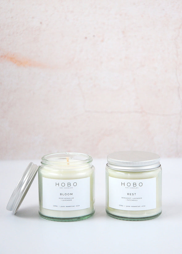 Two aromatherapy travel candles handmade by Hobo Soy Candles, one with its lid on and the other with the lid leaning against its side. Each candle is presented in a one hundred and twenty millilitre recycled glass jar with a metal lid. Each jar is six centimetres high and five point five centimetres wide.