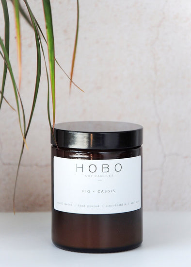 Scented candle in a brown apothecary inspired jar, made by HoBo Soy candles using only natural soy wax. The label shows the scent as being Fig and Cassis. The jar has a capacity of one hundred and eighty millilitres and is eight centimetres tall and six point five centimetres wide.