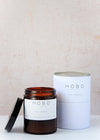 Scented candle in a brown apothecary inspired jar with a black plastic lid, made by HoBo Soy candles using only natural soy wax. Next to the candle is the white packaging which consists of a simple white cardboard cylinder and a metal lid. The label shows the scent as being Fig and Cassis. The jar has a capacity of one hundred and eighty millilitres and is eight centimetres tall and six point five centimetres wide.