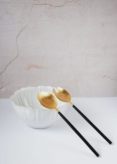 A pair of salad servers made from stainless steel. One has the shape of a spoon and the other the shape of a fork. The handles are black, long and thin while the tops are gold with a width of seven point three centimetres. Each is twenty six centimetres long.