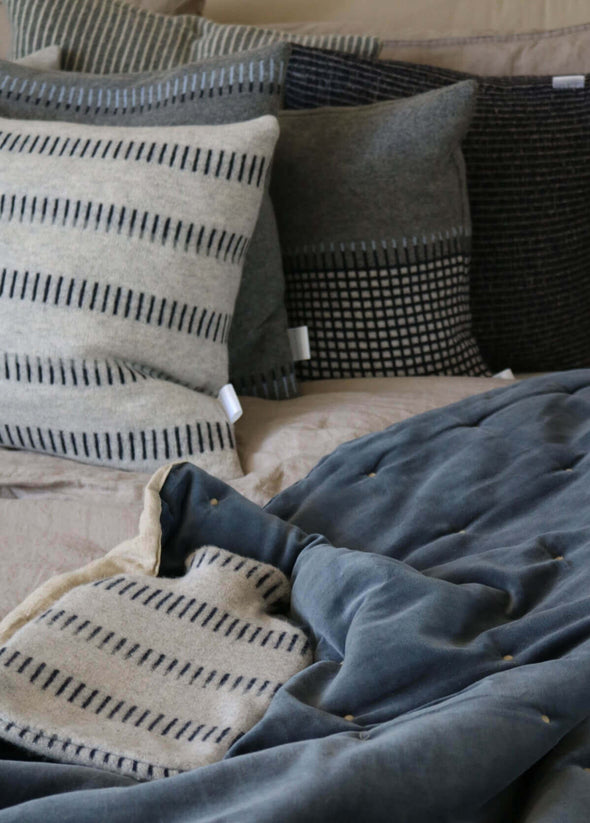 Close up of a bed piled with cushions and a knitted lambswool hot water bottle lying at the front. The hot water bottle is light grey with a navy blue dash pattern.