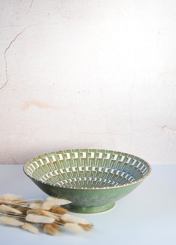 Green bowl with interlocking white design on the inside. Height seven point eight centimetres, width twenty five centimetres. Neutrally coloured pampas grass lies in front of shot.
