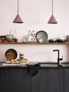 Corner of a kitchen showing a sink in black wooden panelled units with open shelving above. Various kitchenware and tableware items can be seen piled up next to the sink and displayed on the open shelving. Two triangular rose coloured light pendants are hanging just in front of and above the open shelf.
