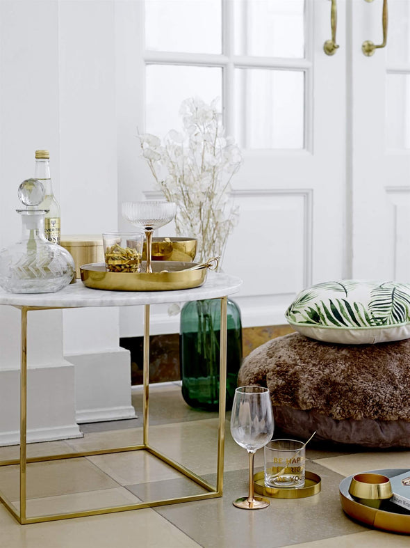 A console table with various glass and gold tableware resting on top. On the floor behind the table is a green glass vase displaying some foliage with pretty white flowers.