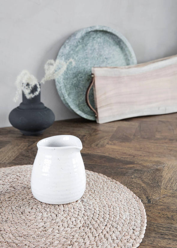 Ivory ceramic milk jug sitting on a weaved placemat. In the background, leaning against a grey wall, are a green marble round tray and wooden rectangular board with a leather handle at the end. A black vase with some white foliage sits next to them.