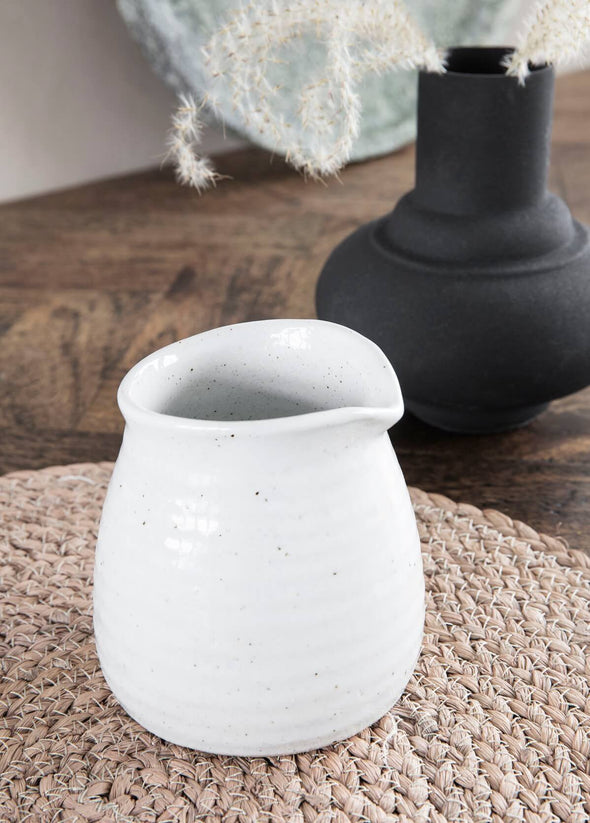 Ivory ceramic milk jug sitting on a weaved placemat. Next to it is a black vase displaying some white foliage.