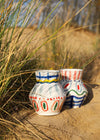 Two ceramic vases set on a sandy beach with grassy dunes as a backdrop. Both vases have a white base and are hand painted with bold brushstrokes using the colours green, orange, blue, yellow and red. They both have a height of fifteen centimetres and a width of twelve centimetres.