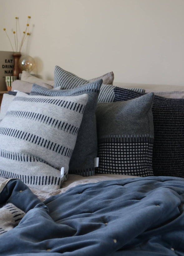Close up of a bed piled with knitted lambswool cushions. The cushions are a mix of grey colours with various blue and white patterns.