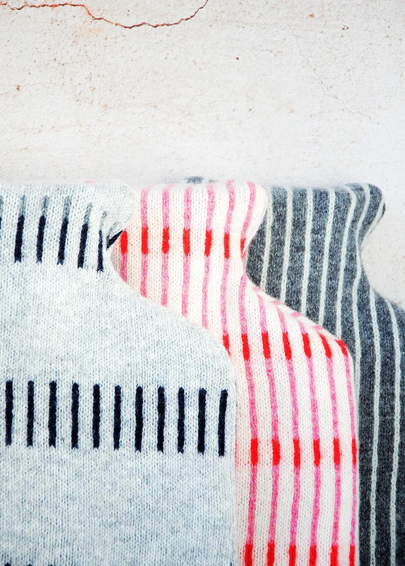 Close up of a group of three knitted lambswool hot water bottles leaning against a wall. The first is light grey with a navy blue dash pattern, the second is white with a pattern of red and pink thin vertical stripes. The third is dark grey with a pattern of white thin vertical stripes.