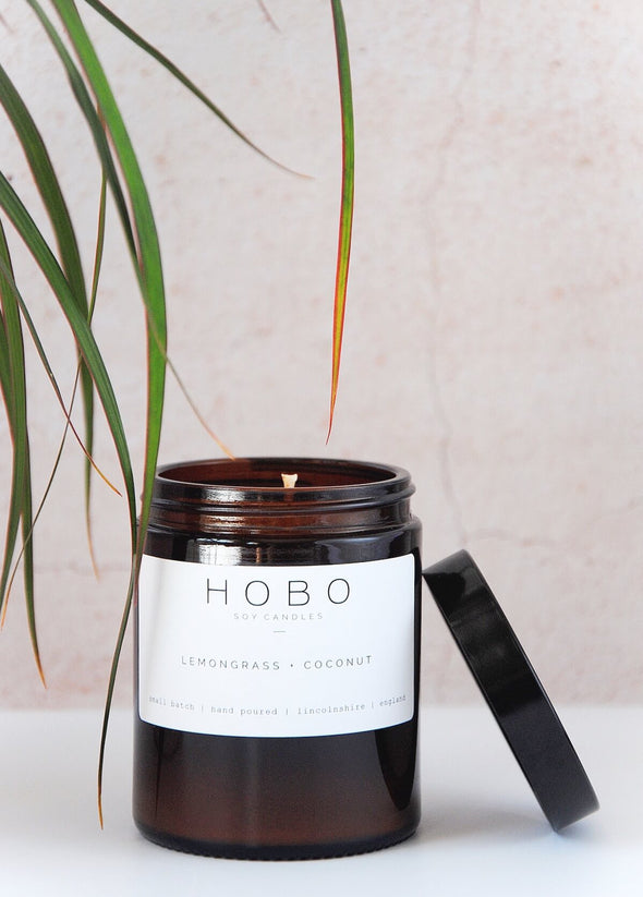 Scented candle in a brown apothecary inspired jar which is open with a black plastic lid leaning against the side, made by HoBo Soy candles using only natural soy wax. The label shows the scent as being Lemongrass and Coconut. The jar has a capacity of one hundred and eighty millilitres and is eight centimetres tall and six point five centimetres wide.