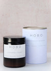  Scented candle in a brown apothecary inspired jar with a black plastic lid, made by HoBo Soy candles using only natural soy wax. Next to the candle is the white packaging which consists of a simple white cardboard cylinder and a metal lid. The label shows the scent as being Lemongrass and Coconut. The jar has a capacity of one hundred and eighty millilitres and is eight centimetres tall and six point five centimetres wide.