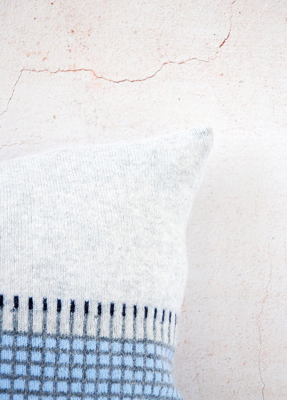 Close up corner of a light grey knitted lambswool cushion with a darker grey and light blue checked pattern on the bottom half. Height forty five centimetres, width forty five centimetres.