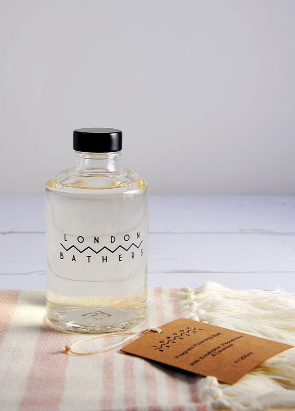 A glass bottle of foam bath by London Bathers sitting on a pink and cream striped cotton towel.