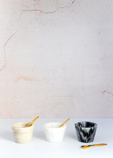 Three marble condiment bowls in a row. A brown marble bowl with brass spoon resting inside, a white marble bowl with brass spoon resting inside and a black marble bowl with brass spoon lying next to it. Each bowl has a height of five centimetres and width of seven point five centimetres.